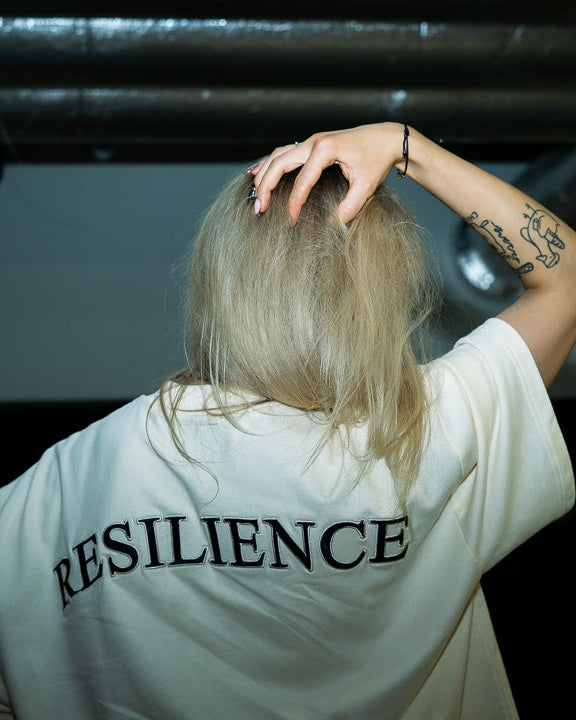A person with blonde hair seen from behind wearing a white oversized T-shirt with the word 'RESILIENCE' printed in black across the back, standing in a dark indoor space with exposed ceiling pipes.