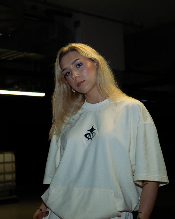 A blonde woman wearing an oversized white t-shirt with a small black graphic on the left chest, standing in a dark, industrial indoor space.