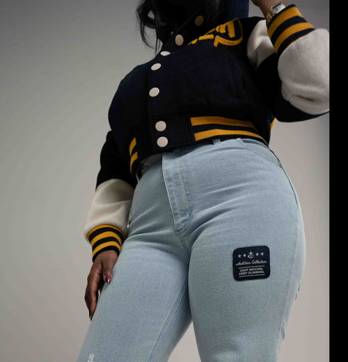 A person wearing a navy and cream varsity-style jacket with yellow stripes, paired with light blue jeans featuring a black patch on the back pocket, against a plain gray studio background.