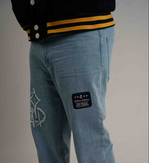 Close-up of a person wearing light blue denim jeans with a black patch on the thigh and a white bicycle graphic on the lower leg, paired with a black varsity-style jacket.
