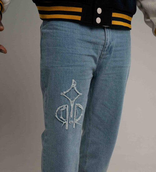 Light blue denim jeans with a white embroidered monogram logo on the left thigh, shown on a model wearing a black and yellow varsity jacket.